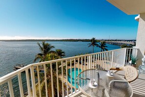 A balcony with a glass table, a chair, and a view of a swimming pool and palm trees.