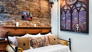 A cozy bedroom with a brick wall, a wooden headboard, a white bed with patterned pillows, a nightstand with a lamp, and a framed picture on the wall.