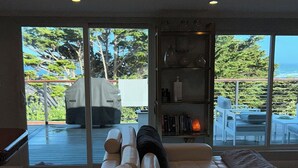 A modern living room with a large sectional sofa, a fireplace, and a view of the outdoors through large windows. - New Home w Expansive Ocean and Sunset Views, Steps to the Seal Cove Beach (Moss Beach)