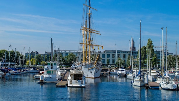 Un puerto deportivo con varios veleros amarrados, un gran barco con múltiples mástiles y edificios al fondo.