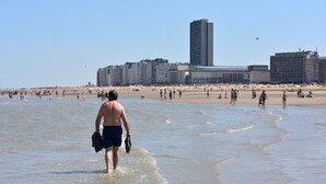 Una playa con gente caminando y de pie, un hombre cargando una chaqueta y zapatos, y edificios al fondo.
