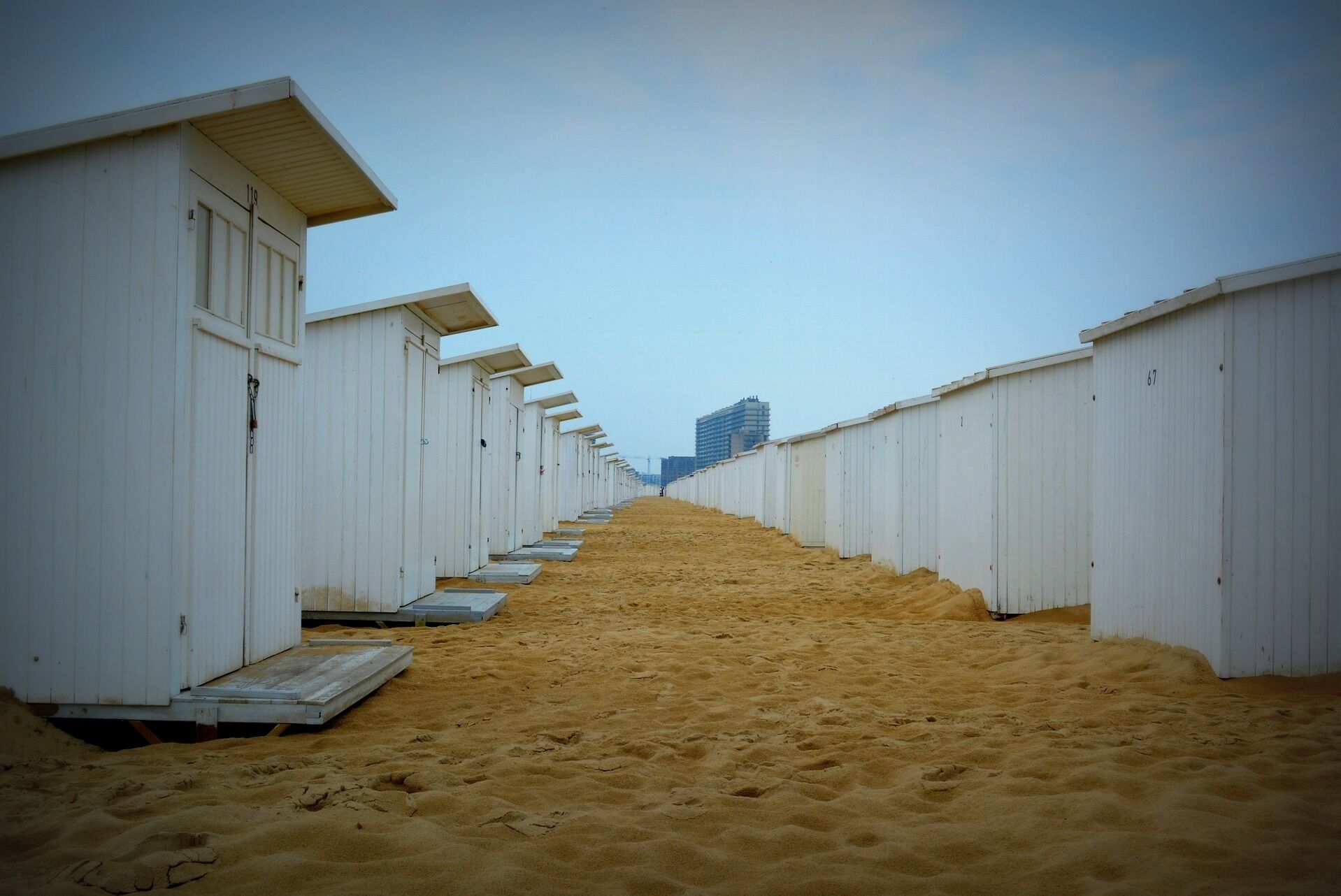 A row of white beach huts with a sandy path leading to a distant building.