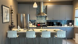 Een moderne keuken met een centraal eiland, barstoelen, roestvrijstalen apparatuur en een hangende lamp.