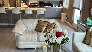 Een moderne woonkamer met een witte leren bank, een glazen salontafel met kaarsen, en een keuken op de achtergrond.