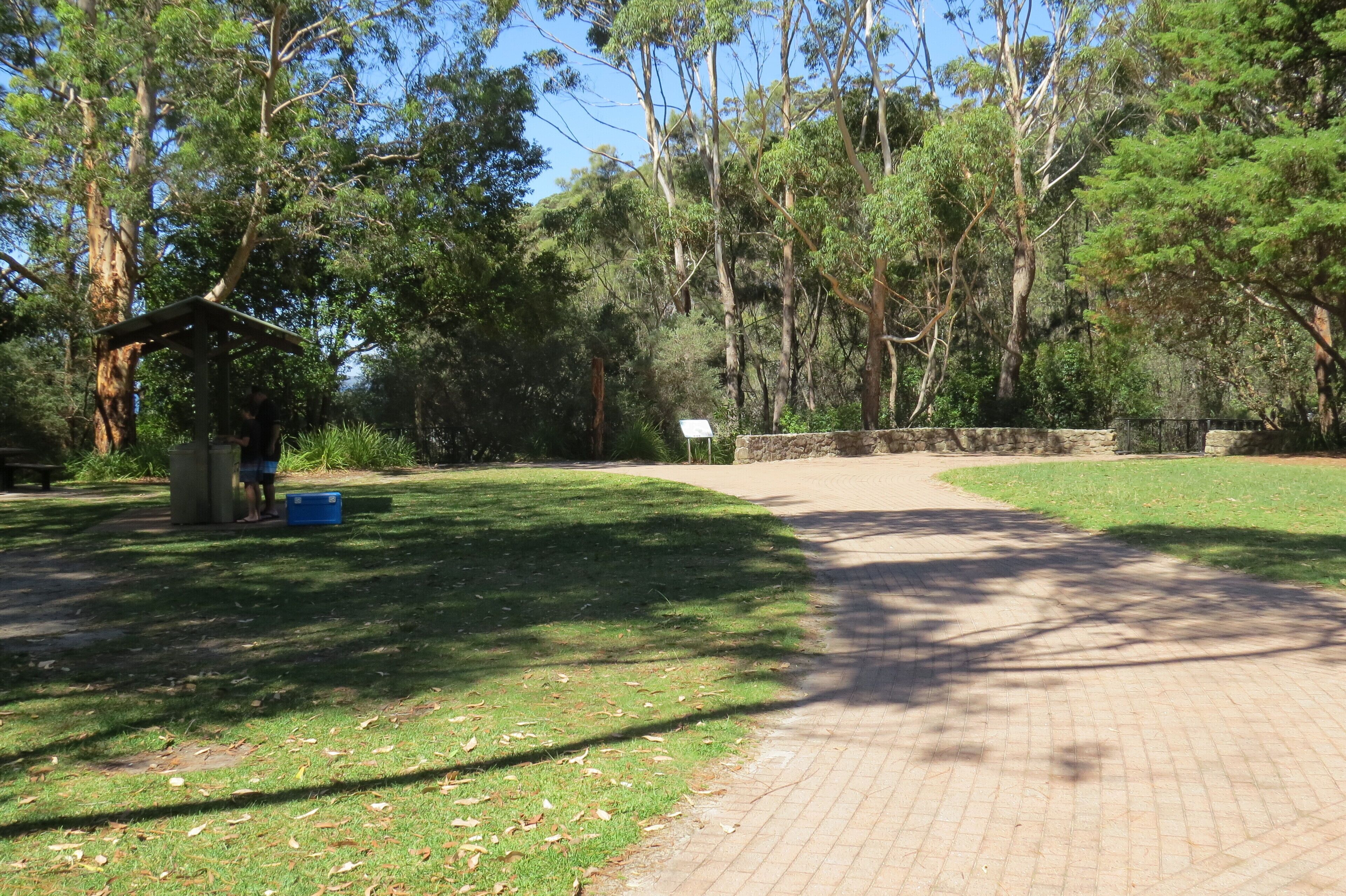 Ein gepflasterter Weg führt durch einen Park mit Bäumen und einer kleinen Schutzhütte.