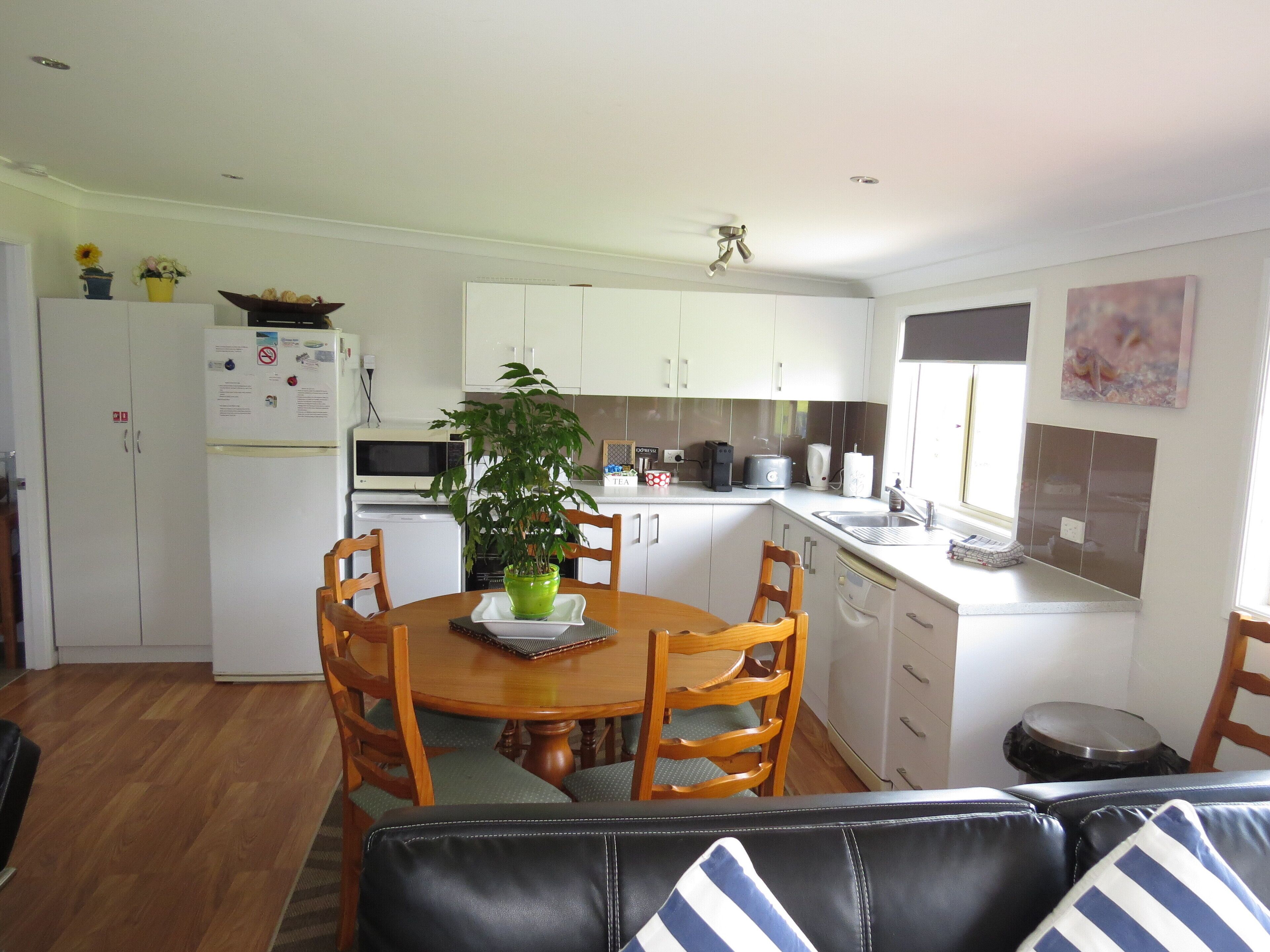 A compact kitchen with white cabinets, a dining table with chairs, a potted plant, and a black leather sofa in the foreground.