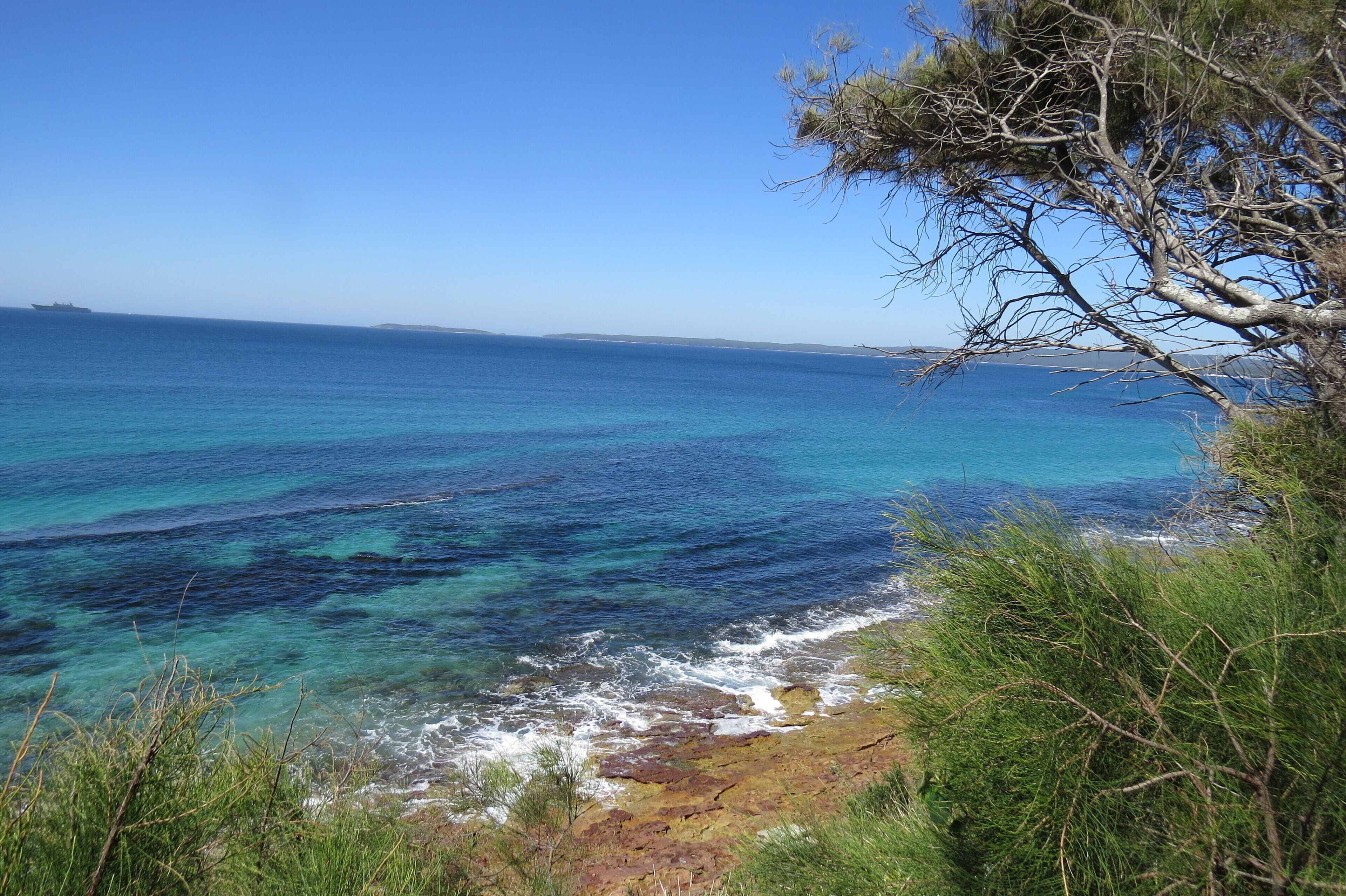 Paisagem costeira com mar azul cristalino, litoral rochoso e vegetação rasteira.