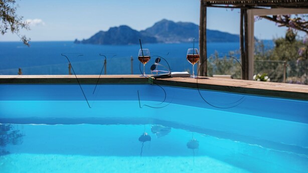 Una piscina con dos copas de vino y gafas de sol en el borde, con vista a una masa de agua y montañas lejanas.
