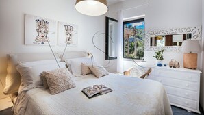 Un dormitorio con cama, almohadas, mesita de noche, lámpara, cómoda y una obra de arte en la pared.