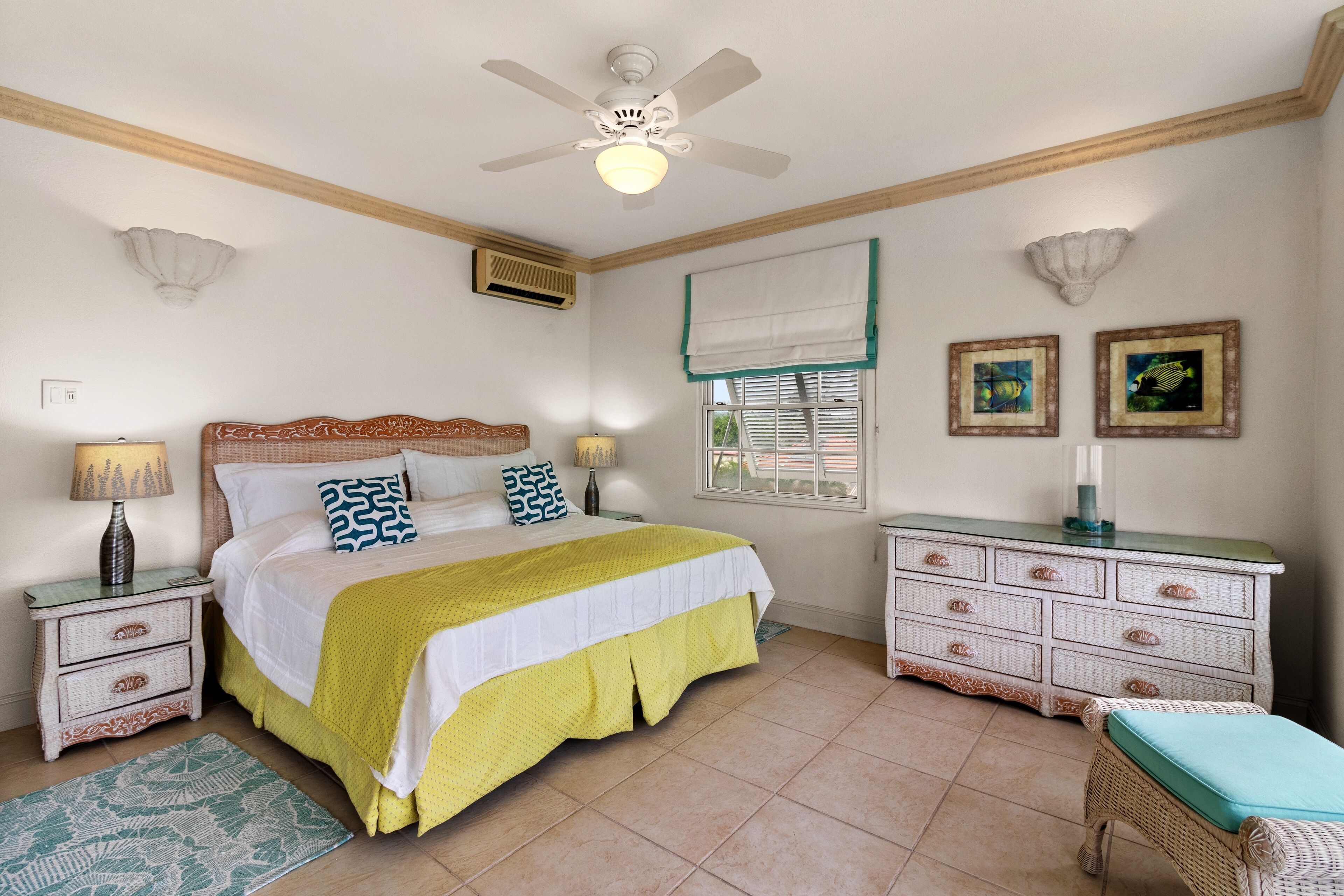 Un dormitorio con cama, mesitas de noche, cómoda, silla, ventilador de techo, ventana con persianas y decoración en la pared.
