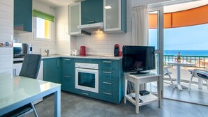 Une cuisine moderne avec des armoires de couleur turquoise, un four encastré et un coin repas comprenant une table et des chaises. Un téléviseur est posé sur une petite table à côté de la cuisine, et un balcon avec vue sur la mer est visible.
