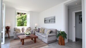 Ein modernes Wohnzimmer mit einem grauen Sofa, gemusterten Kissen, einem Holztisch und einem großen Fenster mit Blick auf Grünflächen.