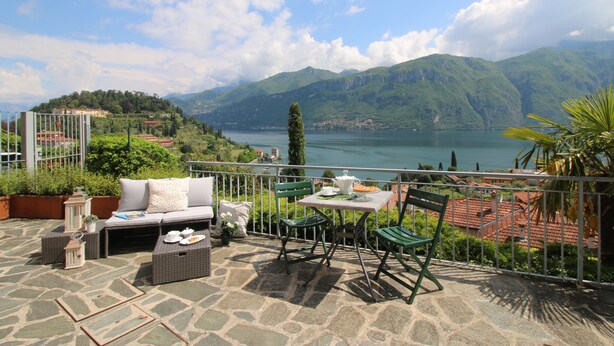 Eine Terrasse mit Steinboden, einer Couch, zwei Stühlen, einem Tisch und Blick auf einen See und Berge.