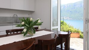 Ein Essbereich mit einem Tisch für vier Personen, einer Vase mit weißen Blumen und Blick auf einen See und Berge durch eine Glastür.