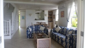 Amplia sala de estar con dos sofás estampados, una mesa de centro de vidrio y una mesa de comedor con sillas. Se observa un ventilador de techo, y una escalera conduce a otro nivel.