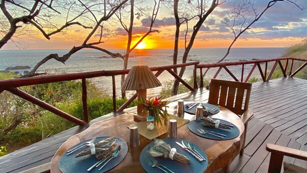 Une terrasse en bois avec une table ronde dressée pour le dîner, offrant une vue sur l’océan au coucher du soleil.