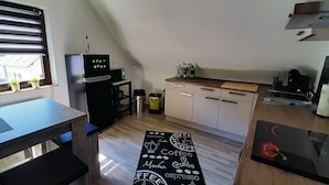 Een kleine keuken met een koelkast, magnetron en koffieapparaat. Op het raamkozijn staan planten in potten en er ligt een koffie-thema vloermat.