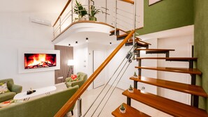 A modern living room with a curved staircase, a flat-screen TV, green sofas, and a small plant on the upper landing.