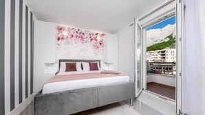 A modern bedroom with a bed, a large window, and a view of buildings and mountains.