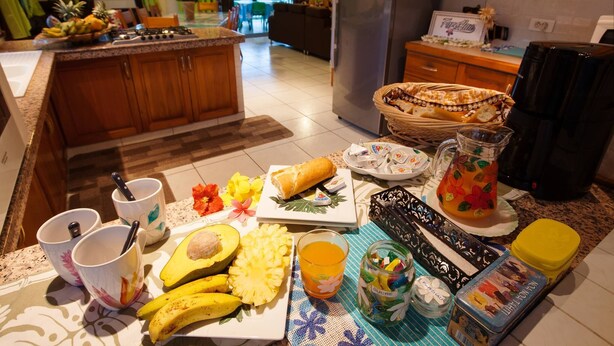 A kitchen with a central island, a dining table with breakfast items, and a refrigerator. - Pension Farehau (Faaa)