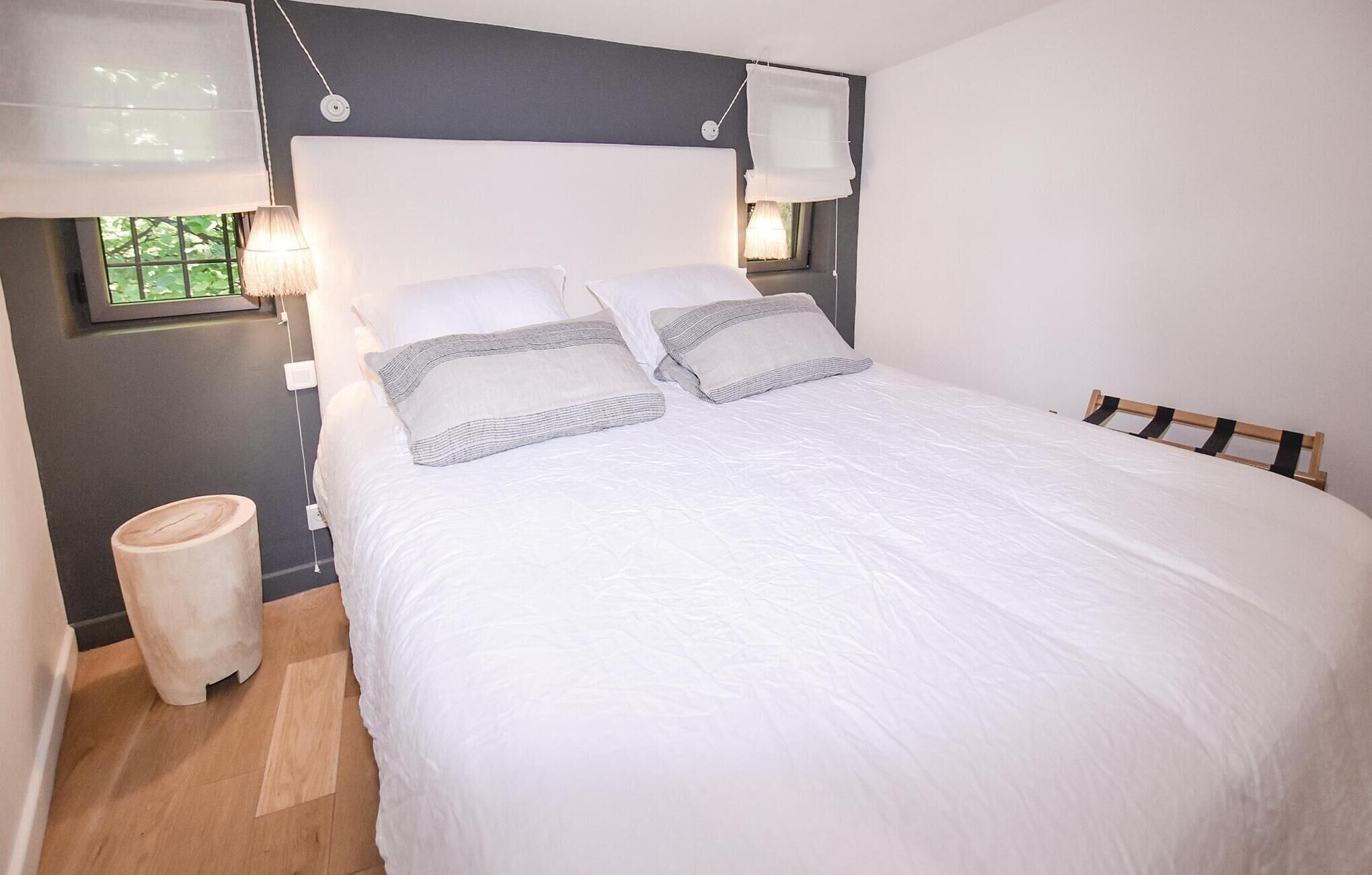 Cama bem arrumada com roupa de cama branca e duas almofadas, luminária de cabeceira e um banquinho de madeira.