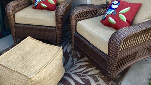 Zwei Korbstühle mit Kissen, ein gewebter Pouf und ein dekoratives Metallobjekt an einer strukturierten Wand.