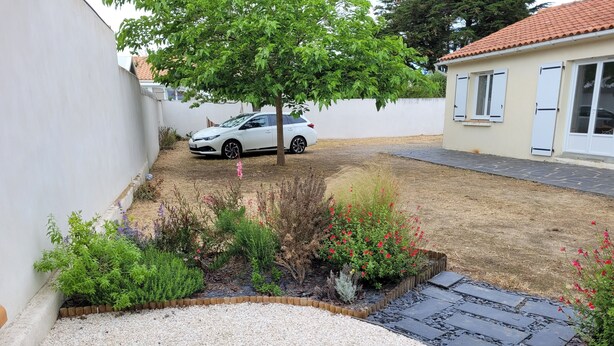 Un barrio residencial con un coche blanco estacionado en una entrada de grava, rodeado de un jardín con diversas plantas y flores.