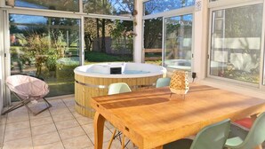 A sunlit patio with a wooden table and chairs, a hot tub, and large windows offering a view of the garden. - Cottage Bien-être Jacuzzi Piscine Chauffée Privée (Peyrole)
