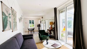 A modern living room with a sofa, a round coffee table, and a green armchair. There is a dining table with chairs in the background, and a kitchen area is visible through an open door. - Cottage Bien-être Jacuzzi Piscine Chauffée Privée (Peyrole)