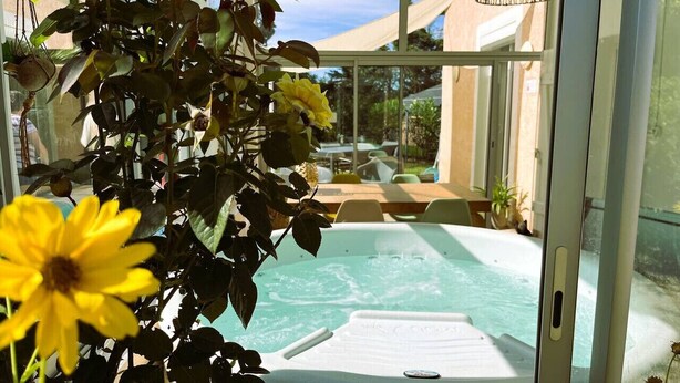A hot tub in a glass-enclosed area with a view of a building and a patio. - Cottage Bien-être Jacuzzi Piscine Chauffée Privée (Peyrole)