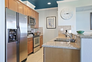 A modern kitchen with stainless steel appliances, wooden cabinets, a microwave, a sink, and a clock on the wall.