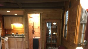 A cozy cabin interior with a wooden floor, a sofa with patterned cushions, a small table, a kitchen area with wooden cabinets, and a window with blinds. - Cozy Cabin at a GREAT price (Whittier)