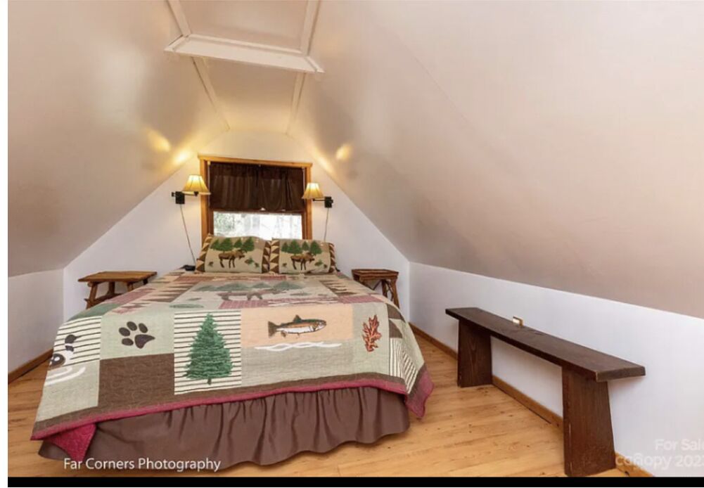 A bedroom with a sloped ceiling, a quilt with animal prints, two bedside lamps, a wooden bench, and a window with curtains.