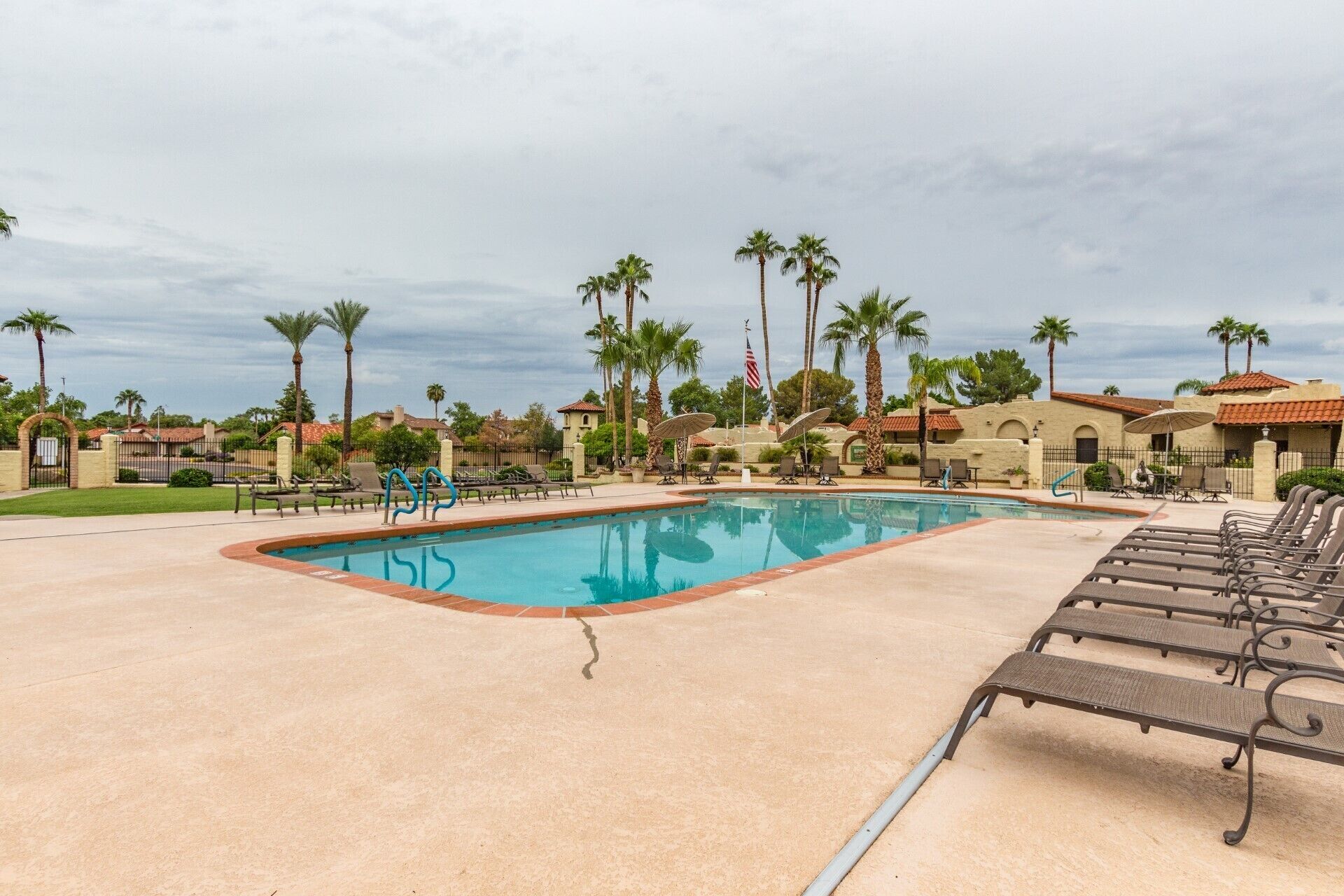 A swimming pool with lounge chairs and umbrellas, surrounded by palm trees and buildings with red-tiled roofs.