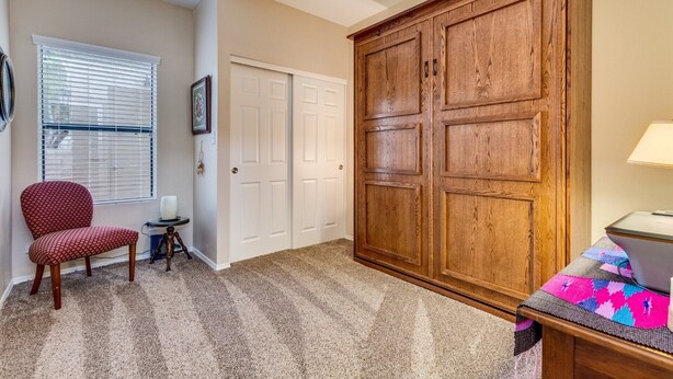 Une pièce comprenant une grande armoire en bois, un fauteuil rouge à motifs, une petite table avec une lampe et une couverture colorée sur un lit.
