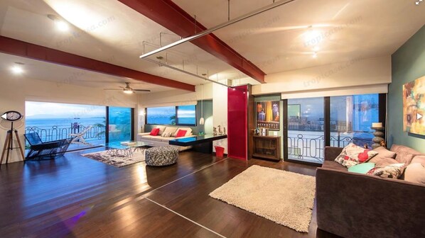 Een ruime woonkamer met een groot raam met uitzicht op zee, een plafondventilator en een gezellige zithoek met een bank en voetenbankje.