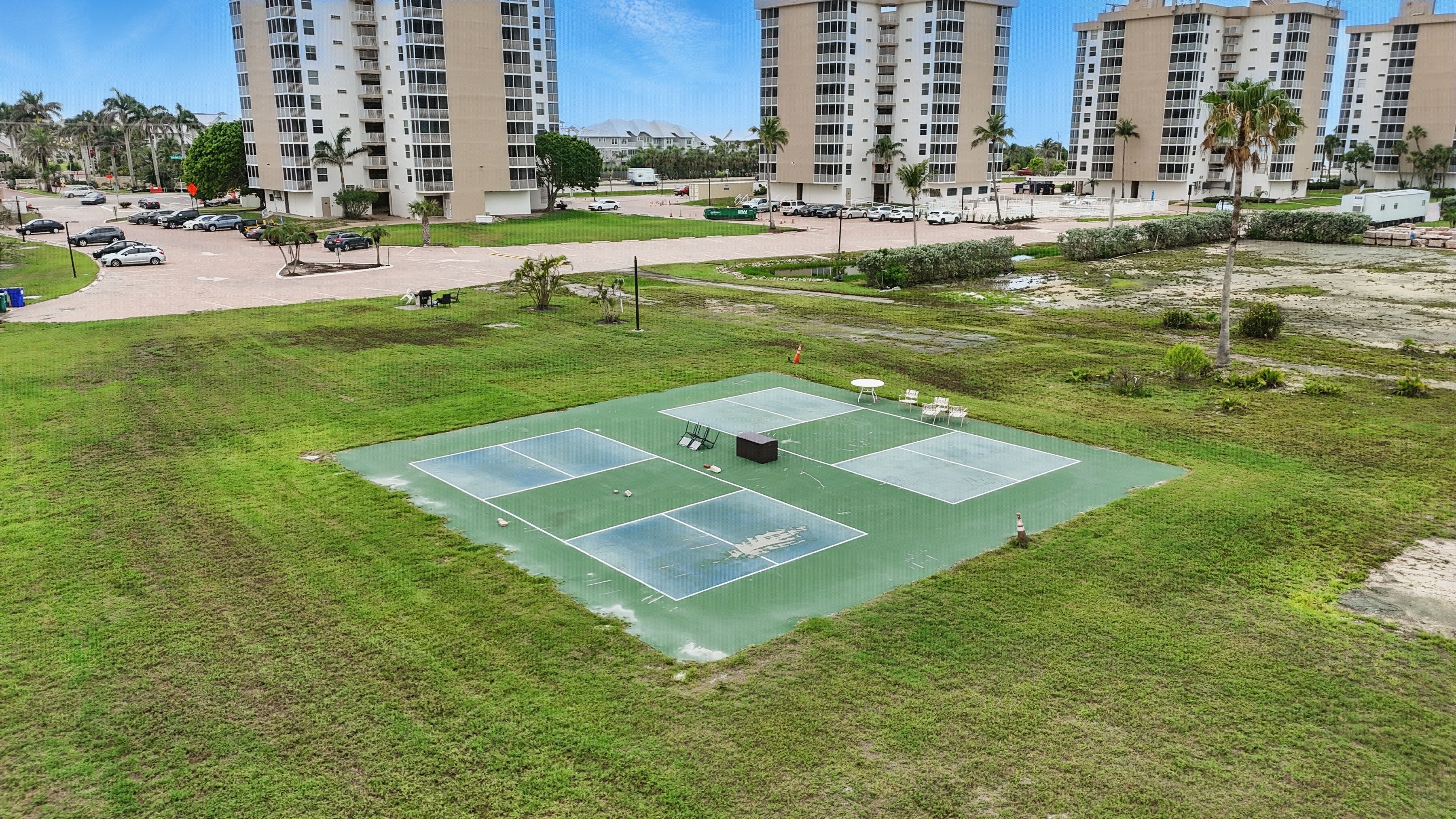 Un court de tennis entouré de pelouse, avec un parking et des bâtiments en arrière-plan.