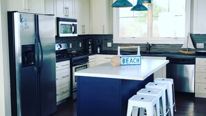 Een moderne keuken met witte kasten, roestvrijstalen apparatuur, een centraal kookeiland met barstoelen, en een bordje met de tekst 'BEACH'.