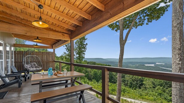 Una terraza de madera con una mesa y sillas de picnic, con vistas a un paisaje pintoresco con árboles y un río.