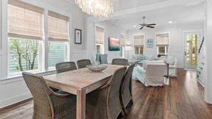 A spacious dining area with a wooden table and wicker chairs, a chandelier, and a view of the outdoors through large windows. - 5 BDRM & 6 seat golf cart! On the green, everything for KIDS, 2 King BDRMs (Santa Rosa Beach)