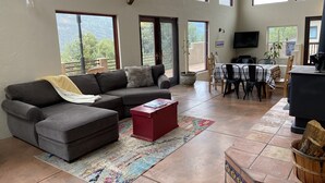 A spacious living room with a gray sectional sofa, a red ottoman, a fireplace, and a dining area with a table and chairs. - Open, Airy Mountaintop Home (Carbondale)