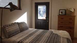 A bedroom with a bed, a desk lamp, a dresser, and a door with a view of trees. - Open, Airy Mountaintop Home (Carbondale)