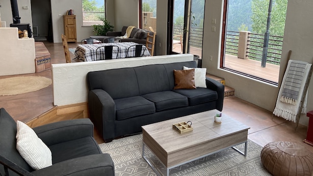 A spacious living area with a black sofa, a wooden coffee table, and a view of the outdoors through large windows. - Open, Airy Mountaintop Home (Carbondale)
