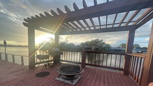 Eine Holzterrasse mit Grill und Feuerstelle mit Blick auf ein Gewässer.