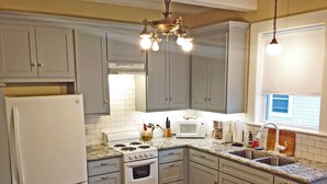 A kitchen with grey cabinets, a white refrigerator, a stove, and a microwave. There is a window with blinds and a chandelier. - Delightful, Relaxing, Restored Historic Apartment in Montrose section of Houston (Houston)