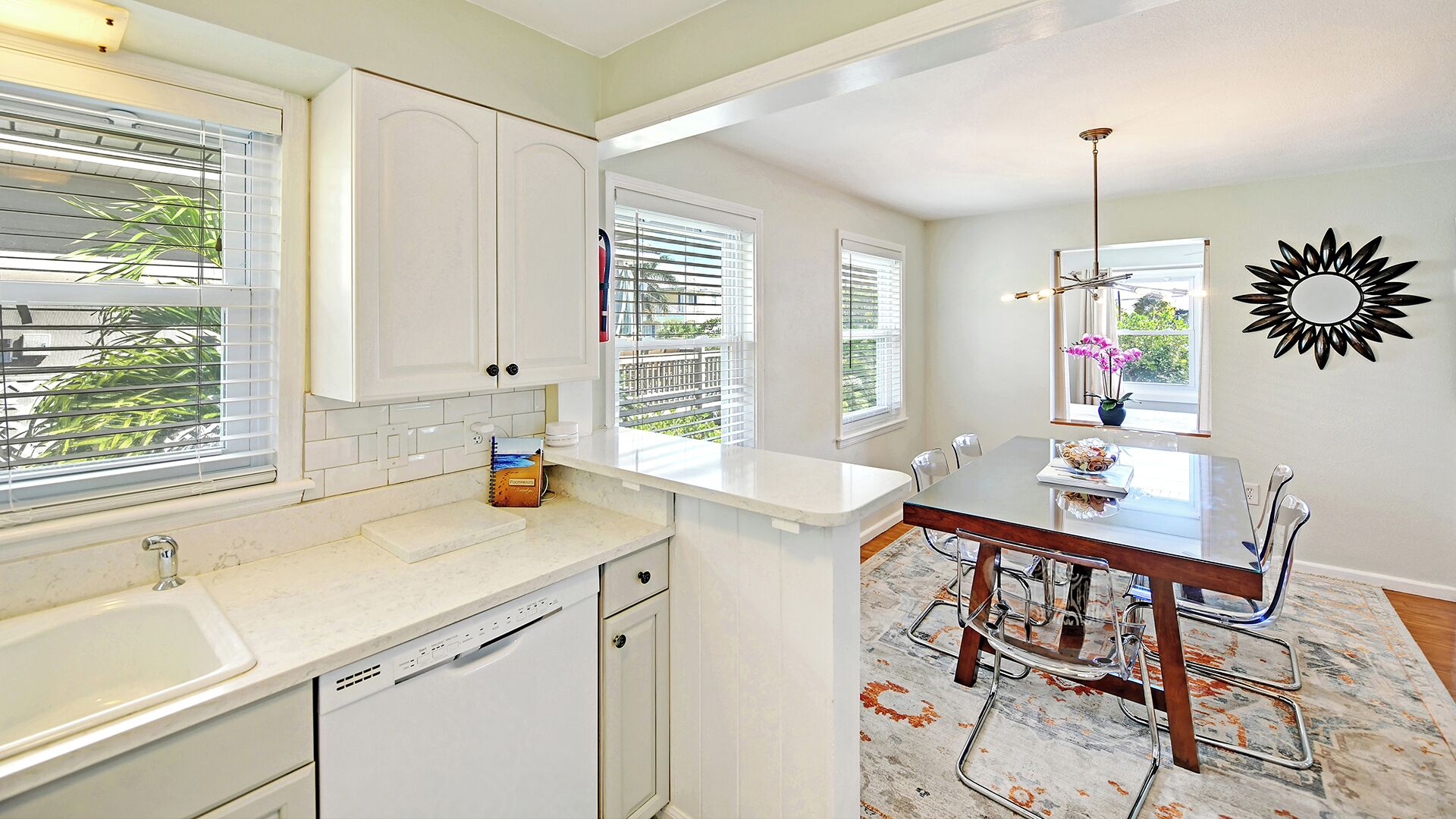 A kitchen with white cabinets, a dishwasher, a dining area with a table and chairs, and a sunburst wall decoration.