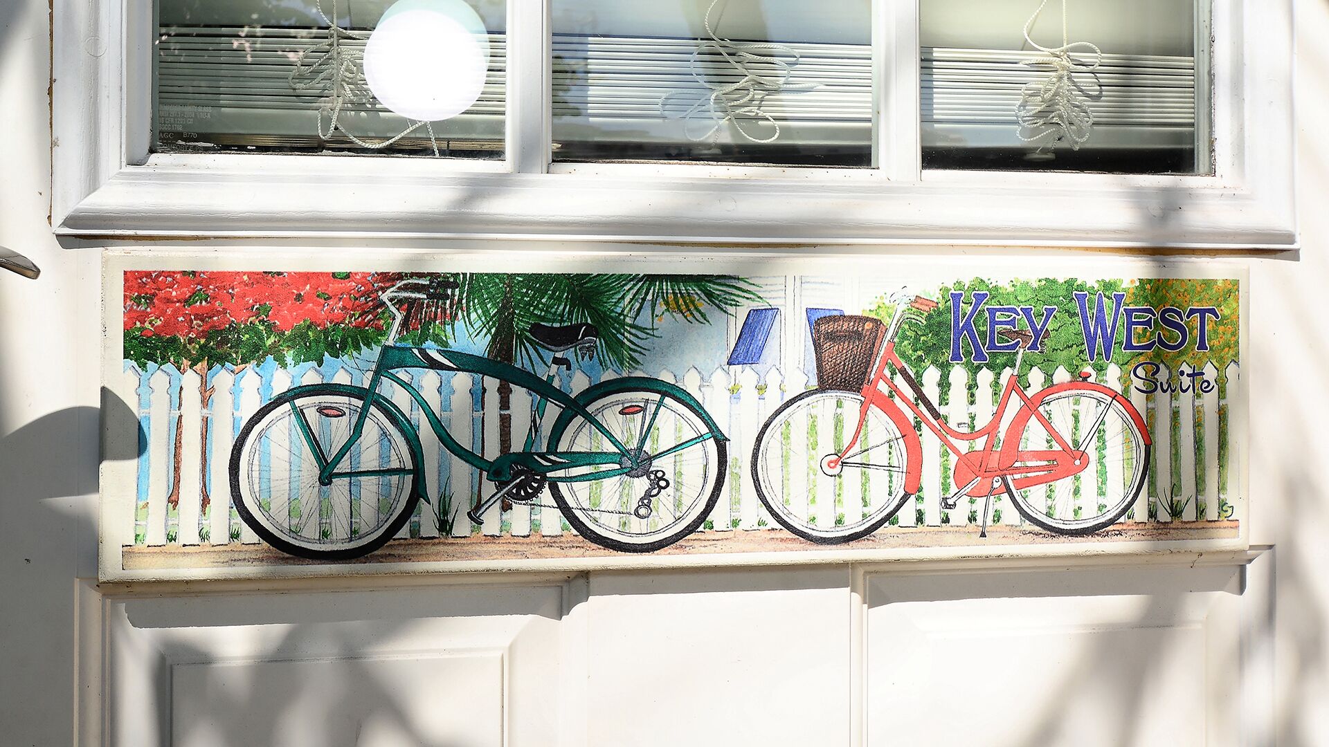 A window with two bicycles painted on it, a white picket fence, and the text "Key West Suites".