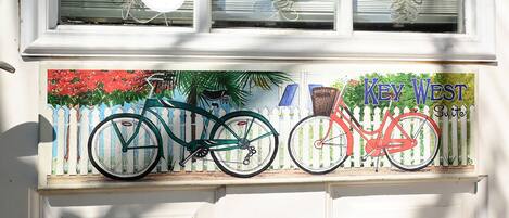 A window with two bicycles painted on it, a white picket fence, and the text "Key West Suites".