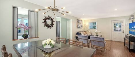 A dining area with a glass table, chairs, and a chandelier. There is a living room with a sofa and a TV.