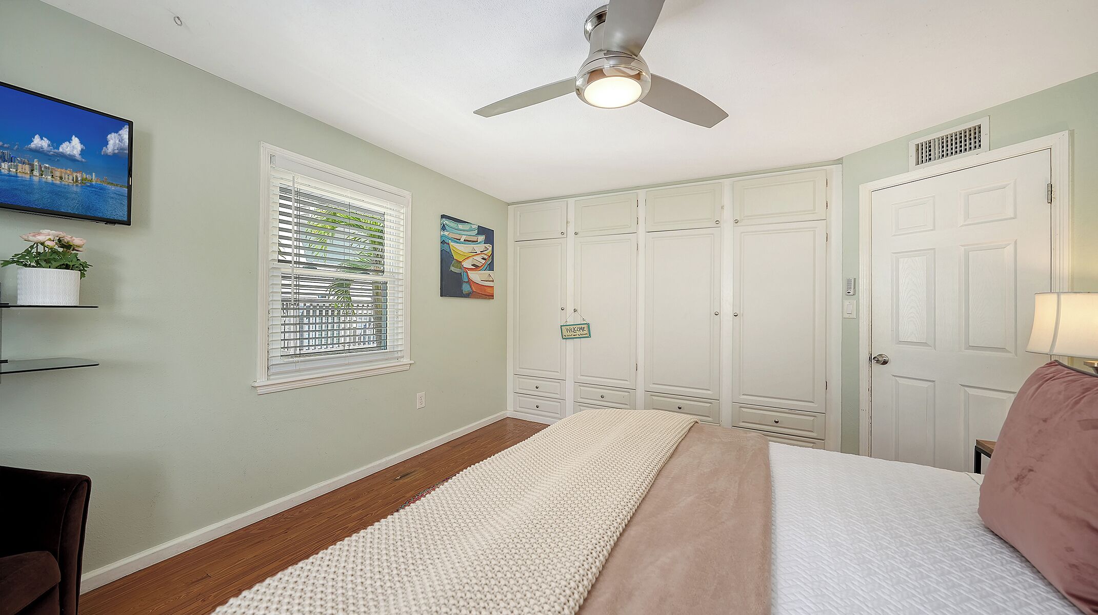 A bedroom with a bed, a ceiling fan, a window with blinds, a welcome sign, and a closet with doors.